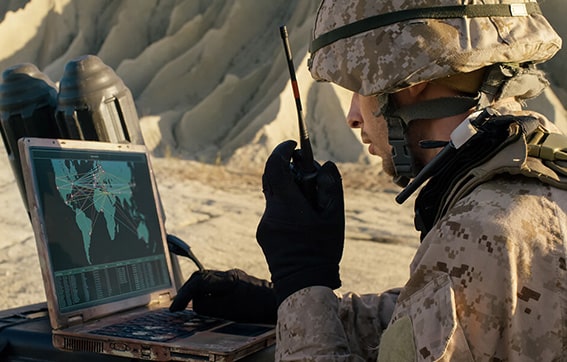 A soldier in camouflage uniform uses a radio and looks at a laptop displaying a world map with network lines, sitting outdoors near rocky terrain.