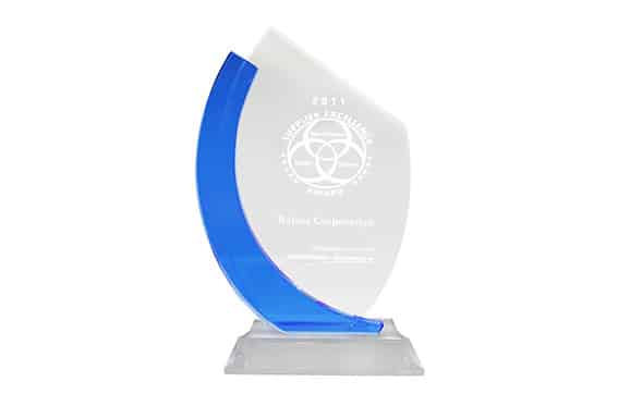 A glass award trophy with a curved blue accent on the left side, mounted on a clear rectangular base. The front features white engraved text and a circular emblem near the top.