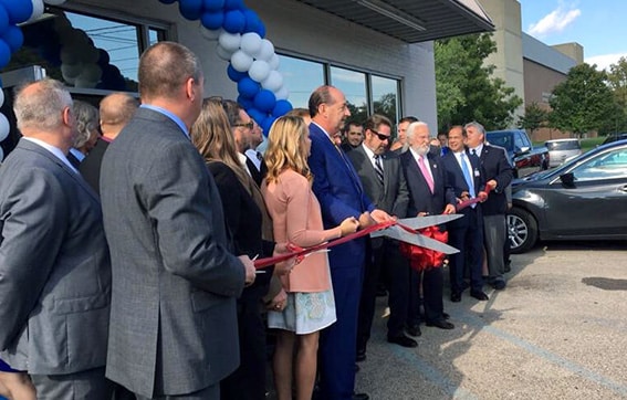 A group of people in formal attire participate in a ribbon-cutting ceremony outside a building decorated with blue and white balloons. Several individuals hold large scissors, preparing to cut a red ribbon.