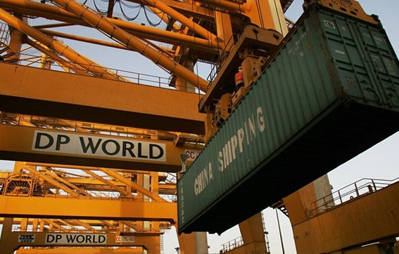 A large green shipping container labeled "China Shipping" is being lifted by a yellow industrial crane at a port operated by DP World. Multiple cranes and metal structures are visible in the background.