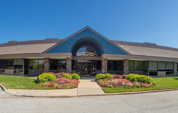 Single-story office building with large windows, stone accents, and an arched entryway. Well-maintained landscaping with green shrubs and pink flowers borders the walkway leading to the entrance under a clear blue sky.