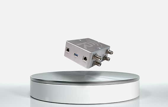 A small metallic electronic device with multiple ports and connectors floats above a round, white display platform against a plain, light background.