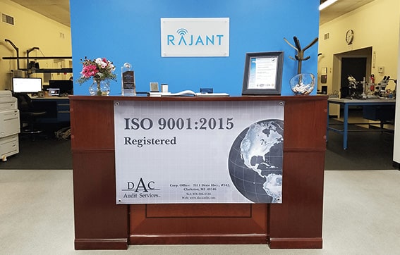 A wooden reception desk displays a large banner reading "ISO 9001:2015 Registered." Behind the desk is a blue wall with a Rajant logo, flowers, a framed certificate, and office equipment visible in the background.