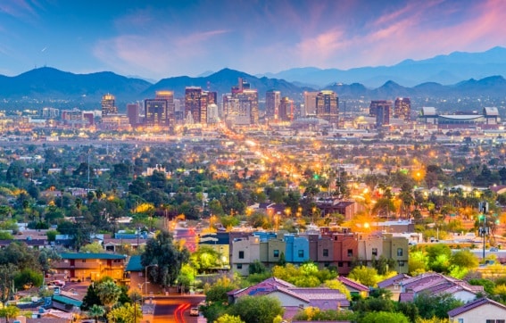 A vibrant cityscape of Phoenix, Arizona, at dusk, with colorful lights illuminating buildings and houses, trees throughout the city, and mountains visible in the background under a dramatic sky.
