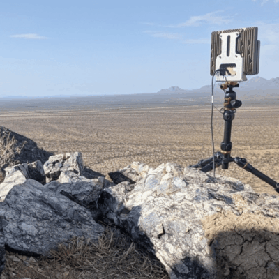 A portable satellite communication device mounted on a tripod stands on rocky terrain overlooking a vast, arid desert landscape with distant mountains under a clear sky.