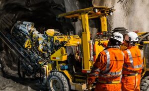 Two workers in orange safety gear and helmets operate a large yellow drilling machine inside an underground mine tunnel with rough rock walls.