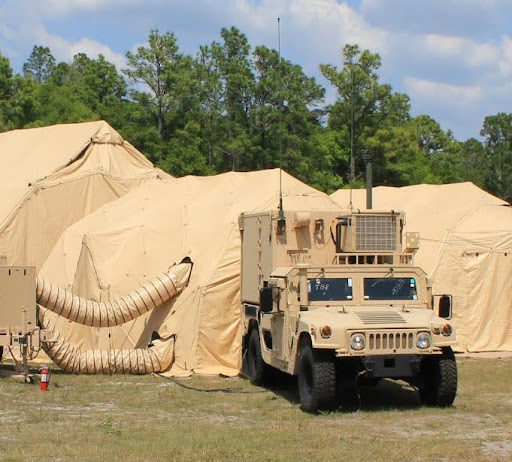 A beige military vehicle is parked in front of large tan tents with ventilation hoses, set up outdoors on grass with trees and a blue sky in the background.