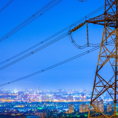 A large electrical transmission tower with power lines stands in the foreground, overlooking a brightly lit cityscape at dusk with a clear blue sky.