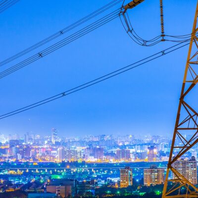 A large electrical transmission tower stands in the foreground at dusk, with power lines stretching across the image. In the background, a brightly lit cityscape glows under a blue evening sky.