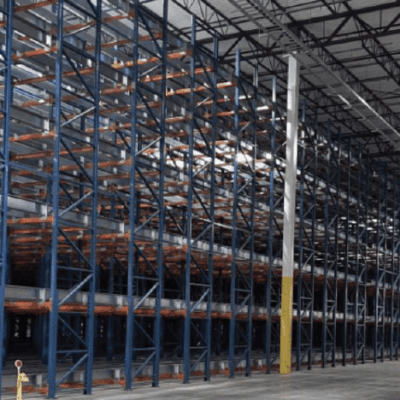 A large, empty warehouse interior with tall, blue and orange metal storage racks lined up in rows beneath a high ceiling with exposed beams. The concrete floor is clean and unobstructed.