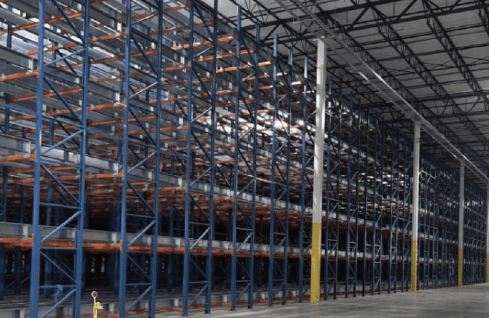 A large, empty warehouse interior with tall, blue and orange metal storage racks lined up in rows beneath a high ceiling with exposed beams. The concrete floor is clean and unobstructed.
