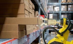 A robotic arm retrieves a cardboard box from a shelf in a warehouse stocked with rows of similar boxes.