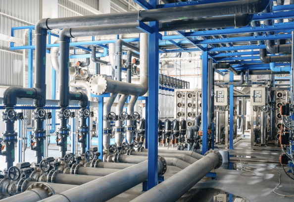 Interior of an industrial facility featuring complex interconnected pipes, valves, control panels, and metal structures, likely part of a water treatment or processing plant, all in a clean, well-lit environment.