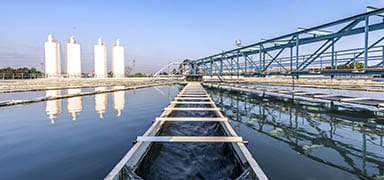 A large water treatment facility with clear water pools, metal walkways, and several tall white cylindrical tanks in the background under a blue sky.