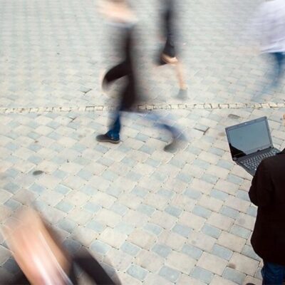 A man in a black jacket stands on a cobblestone street using a laptop, while blurred figures of people walk quickly around him, suggesting movement and busyness.