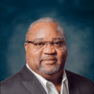 A middle-aged man with short gray hair, glasses, and a trimmed beard, wearing a dark suit jacket and white shirt, posed against a blue-gray studio background.