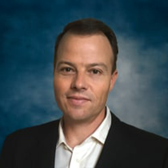 A man with short brown hair wearing a black blazer and white collared shirt, posing against a blue, softly blurred background.