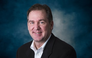 A middle-aged man with short brown hair, wearing a white shirt and dark blazer, smiling at the camera against a blue-gray studio background.