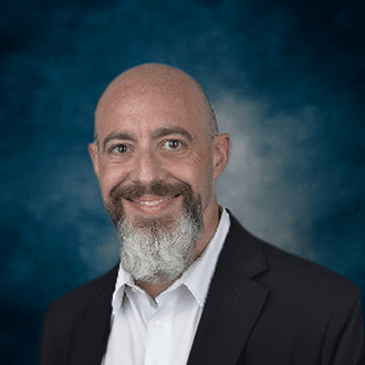 A smiling bald man with a gray and white beard wearing a dark suit jacket and white shirt, posed against a blue and gray mottled background.