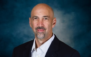 A middle-aged man with a bald head and goatee wearing a dark suit jacket and white collared shirt poses in front of a blue mottled background.