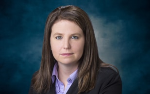 A woman with straight brown hair, wearing a dark blazer over a lavender collared shirt, poses for a formal portrait against a blue, softly blurred background. She has a neutral expression.