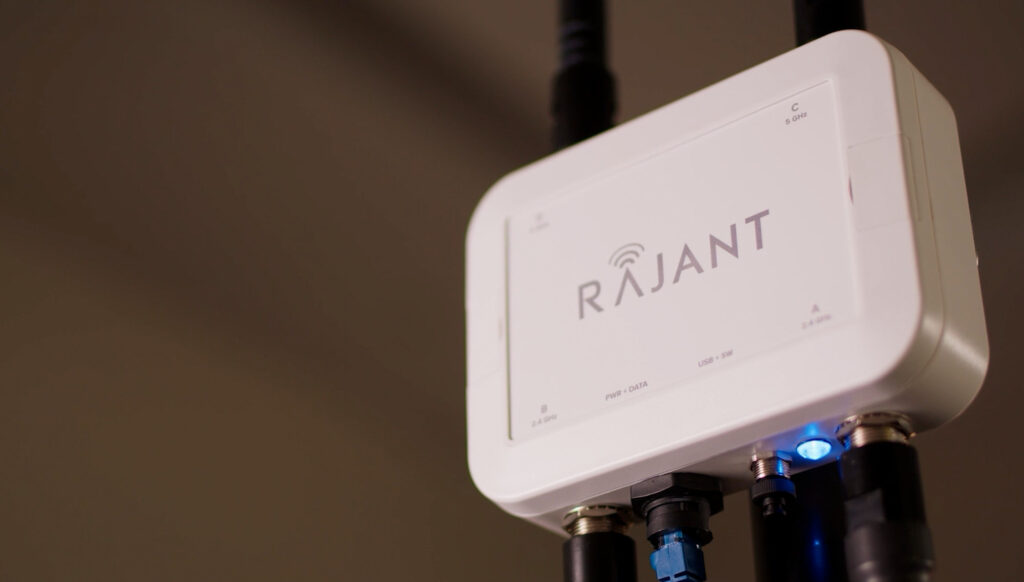 Close-up view of a white Rajant wireless networking device mounted with several black cables connected to it, and a blue LED indicator light glowing.
