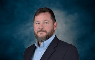 A man with short dark hair and a beard, wearing a dark blazer and light blue collared shirt, poses against a blue, cloudy backdrop.
