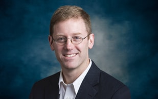A man with short light brown hair and glasses, wearing a dark suit jacket and white collared shirt, smiles in front of a blue and gray mottled background.