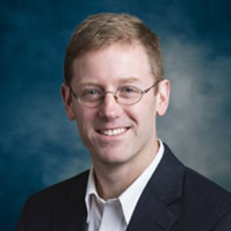 A man with short light brown hair, wearing glasses, a white shirt, and a dark suit jacket, smiles in front of a blue background.