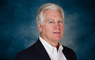A middle-aged man with short white hair is wearing a dark suit jacket and white collared shirt, posing against a blue textured background.