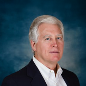 A middle-aged man with short gray hair wearing a dark blazer and white shirt poses in front of a blue background.