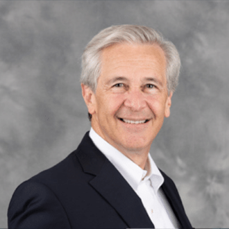 A smiling older man with gray hair, wearing a dark suit jacket and white shirt, poses in front of a gray, cloudy backdrop.