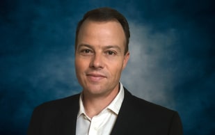 A man with short brown hair wearing a black suit jacket and white shirt poses in front of a blue, cloudy background.