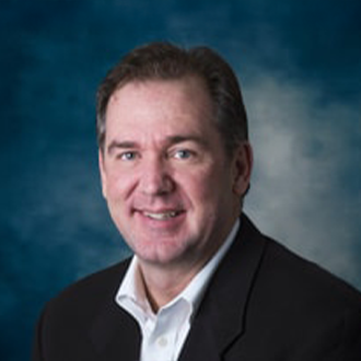 A middle-aged man with short brown hair, wearing a dark suit jacket and white collared shirt, smiles at the camera against a blue and gray gradient background.