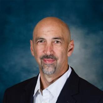 A middle-aged man with a bald head and goatee is wearing a dark suit jacket and white shirt, posing in front of a blue and gray mottled background.