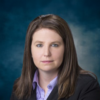 A woman with straight brown hair wearing a black blazer over a light purple collared shirt poses in front of a blue and gray studio background.