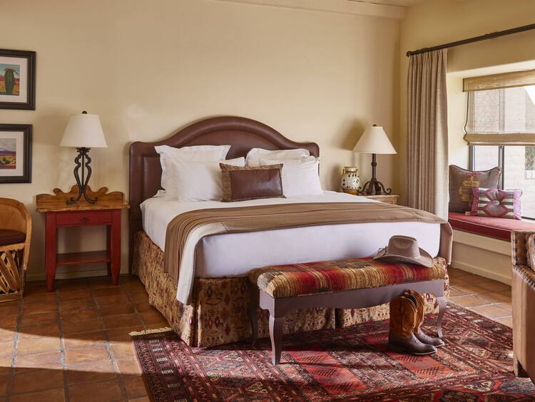 A cozy bedroom with a large bed, patterned bench, southwestern-style rug, bedside tables with lamps, cowboy boots and hat on the floor, and a window seat with cushions and natural light.
