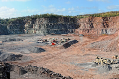A wide panoramic view of an open-pit quarry with terraced rock walls and scattered mining equipment on the dusty floor, surrounded by trees and under a partly cloudy sky.