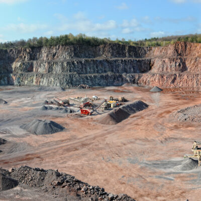 A wide panoramic view of an open-pit quarry with terraced rock walls and scattered mining equipment on the dusty floor, surrounded by trees and under a partly cloudy sky.