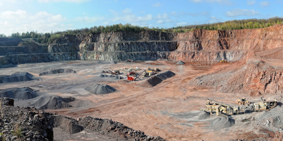 A wide panoramic view of an open-pit quarry with terraced rock walls and scattered mining equipment on the dusty floor, surrounded by trees and under a partly cloudy sky.