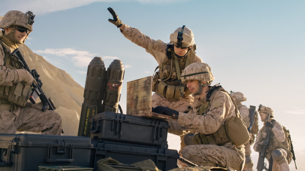 Three soldiers in desert camouflage gear operate military equipment outdoors. One gestures with an arm raised, while another examines a device on a case. Rugged terrain and blue sky are visible in the background.