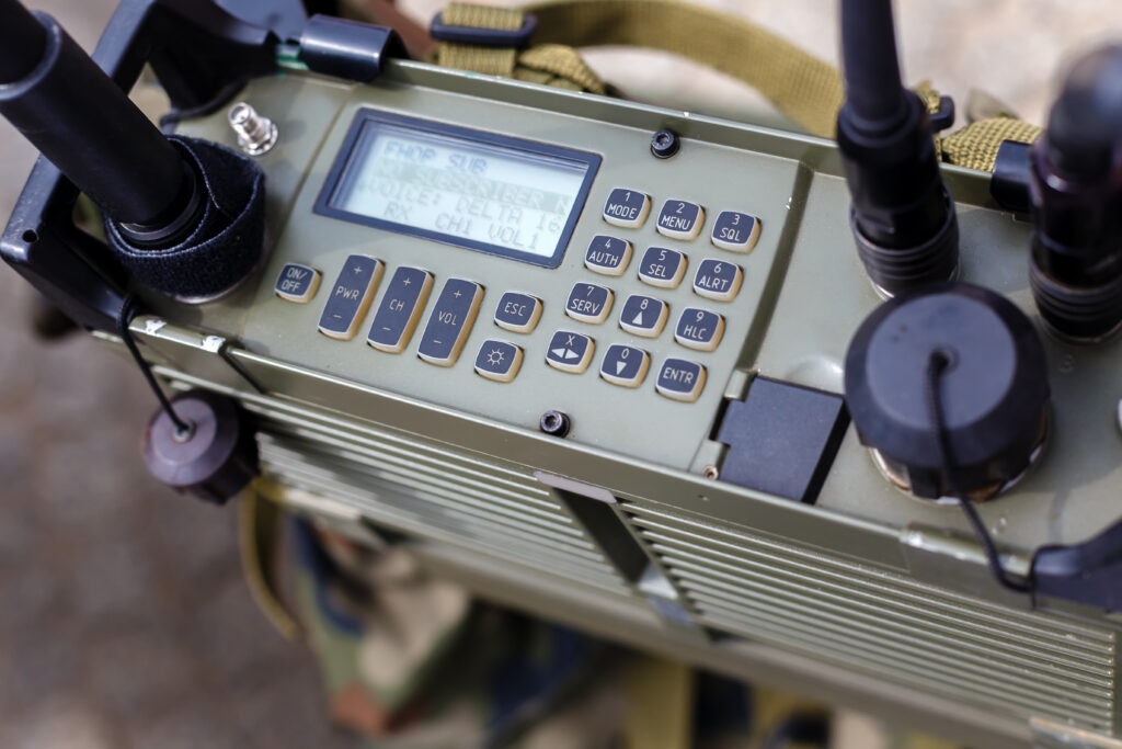 Close-up of a military radio communication device with a digital display, several buttons, knobs, and connected cables. The screen shows signal and frequency information.
