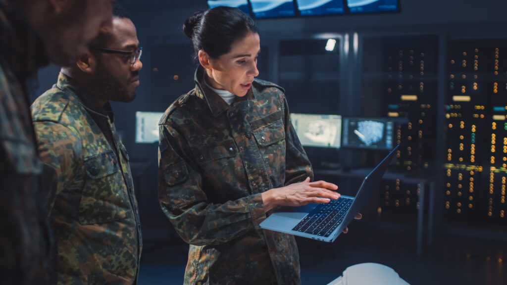 Three military personnel in camouflage uniforms stand in a high-tech control room; one woman is holding a laptop and showing something on the screen to her colleagues. Multiple monitors and servers are visible in the background.