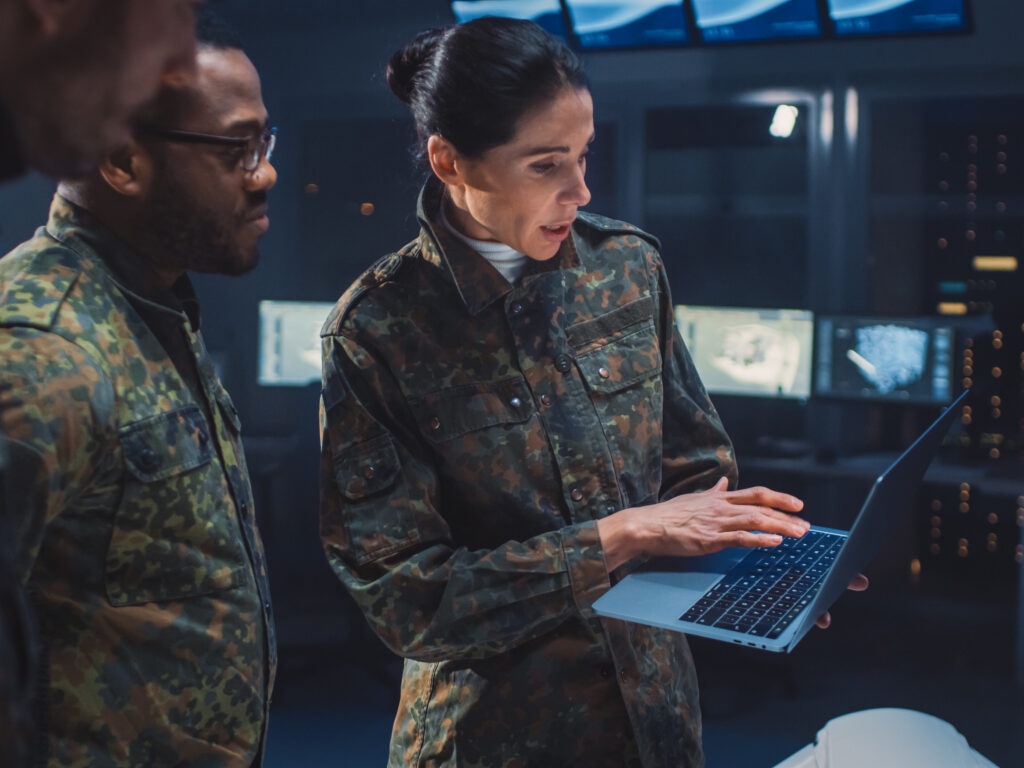 Three military personnel in camouflage uniforms stand in a high-tech control room; one woman is holding a laptop and showing something on the screen to her colleagues. Multiple monitors and servers are visible in the background.