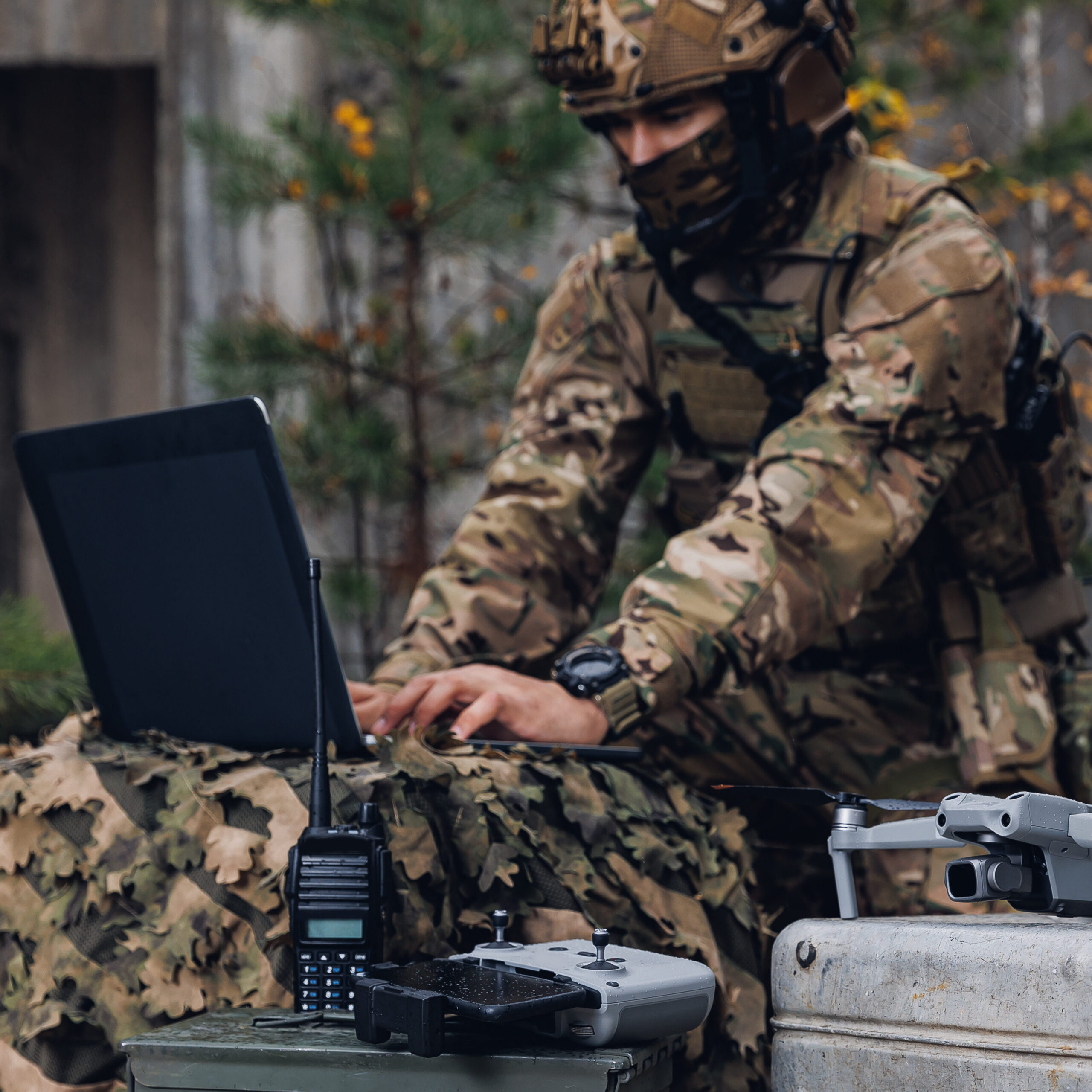 A soldier in camouflage gear works on a laptop outdoors, surrounded by equipment including a drone, a walkie-talkie, and other military gear. Autumn leaves and a concrete structure are visible in the background.