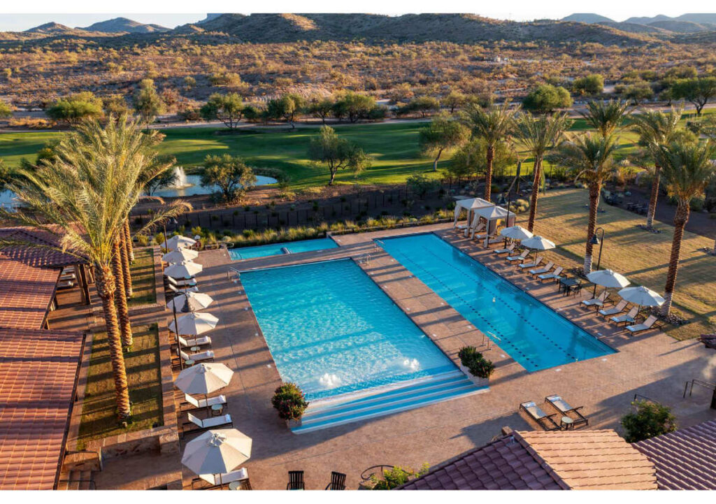 Aerial view of a large outdoor swimming pool with lounge chairs and umbrellas, surrounded by palm trees, lawns, and desert landscape with mountains in the background.