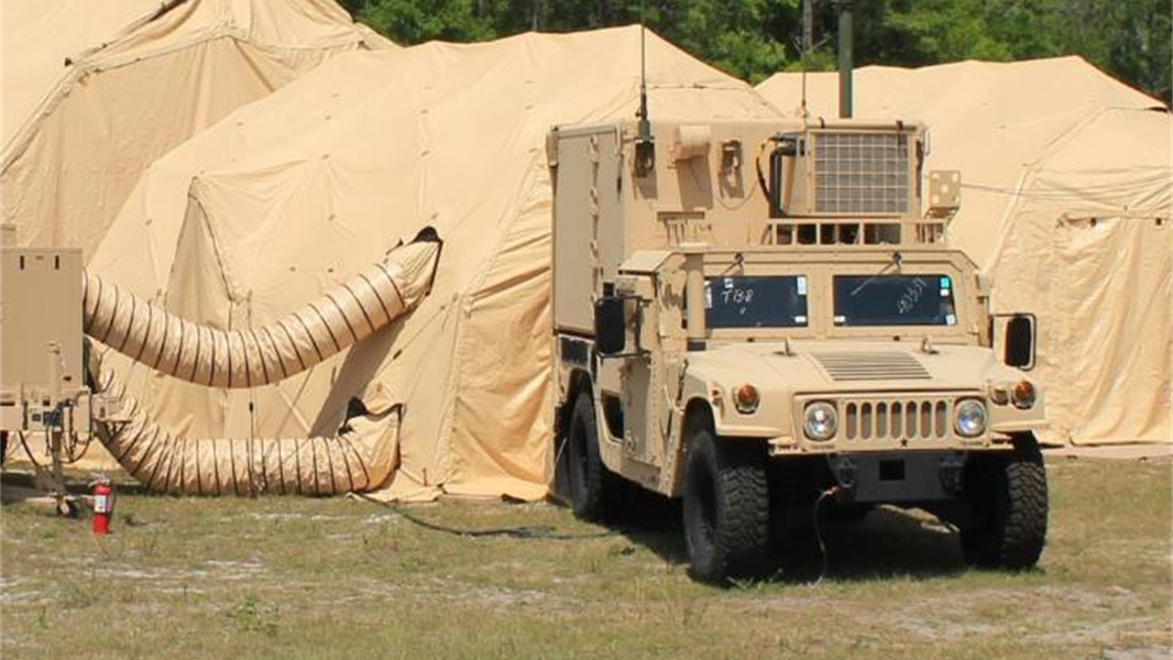 A tan military Humvee is parked on grass beside large beige tents with ventilation ducts, set against a backdrop of trees and blue sky.
