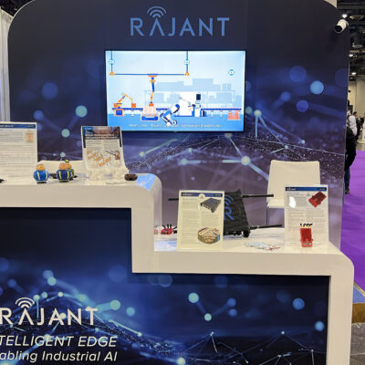 A trade show booth for Rajant displays technology solutions with a large screen, informational materials, and device demos. Visitors are walking by on a purple-carpeted floor in a busy exhibition hall.
