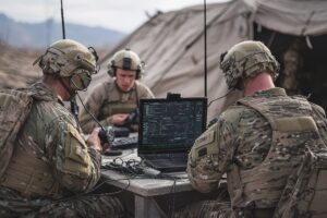 Three soldiers in camouflage uniforms and helmets operate communication equipment and a laptop with tactical maps and data, sitting at a table outside a military tent in a rugged, mountainous area.