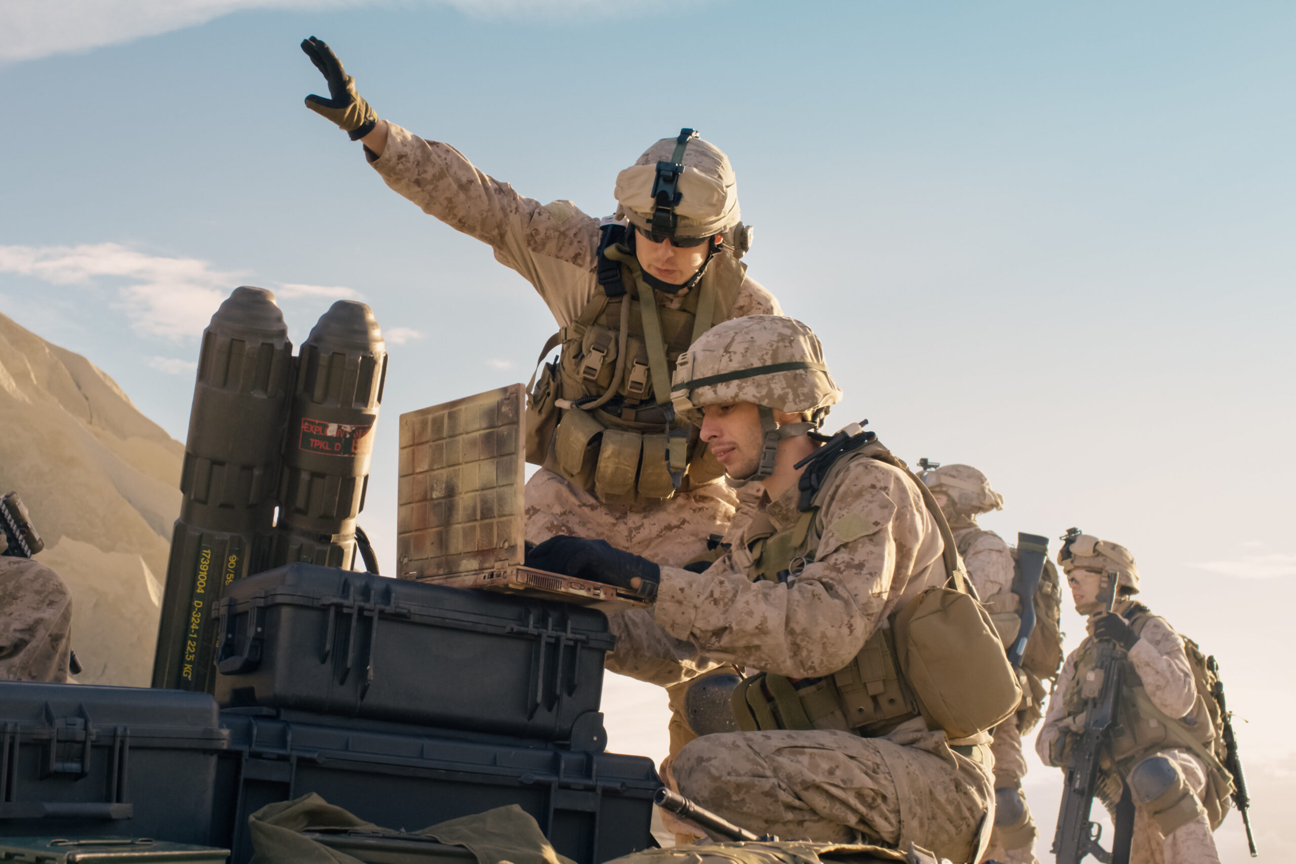 Three soldiers in desert camouflage gear operate military equipment outdoors. One gestures with an arm raised, while another examines a device on a case. Rugged terrain and blue sky are visible in the background.
